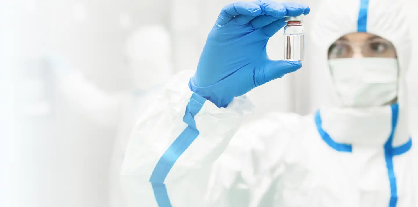 A doctor wearing goggles, a face mask, a protective suit, and plastic gloves holds a small jar of liquid in front of their face.