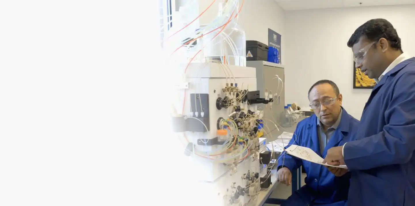 Two men in blue lab coats and goggles look at a document that one of them is holding next to YMC equipment.